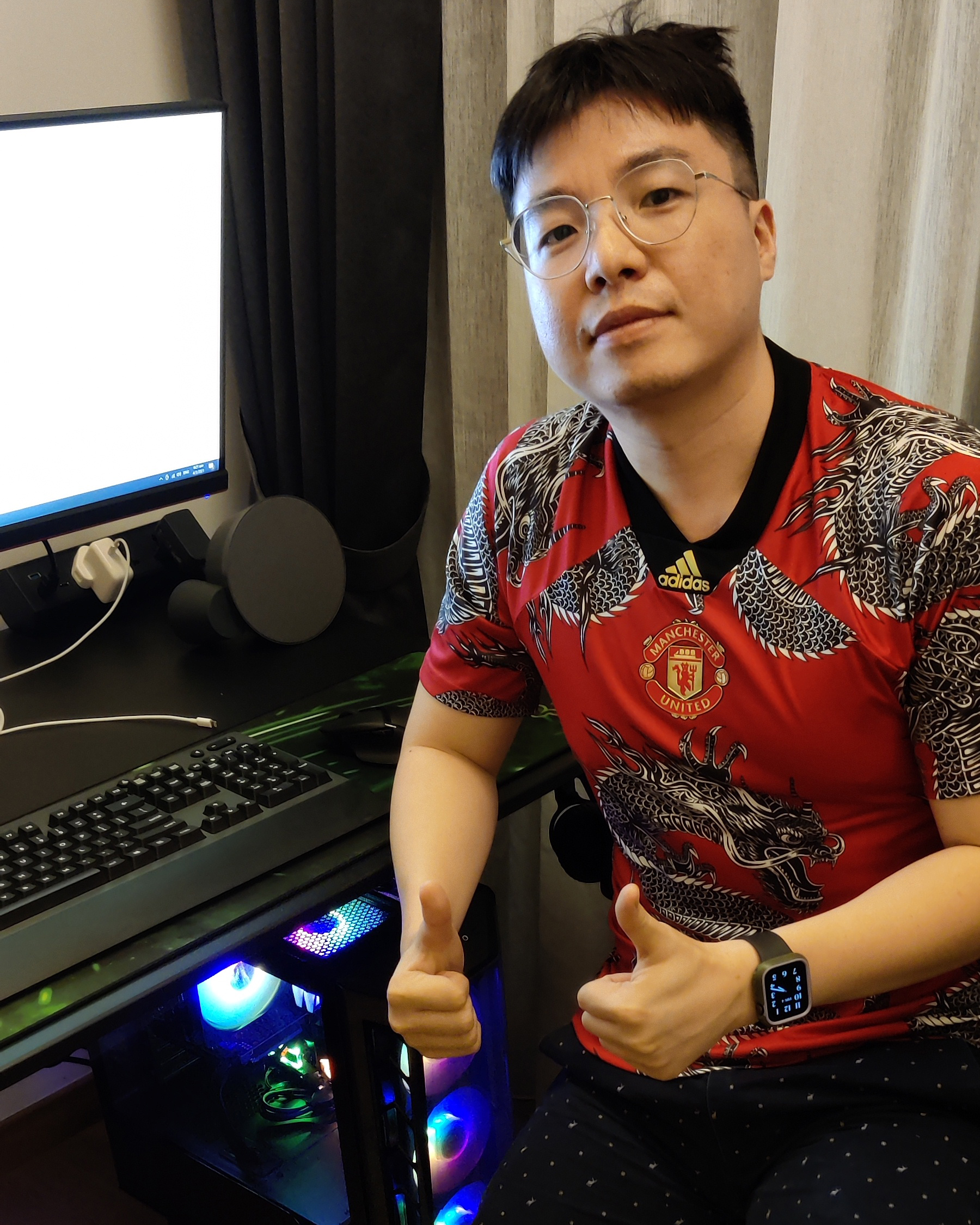 Satisfied PCExperts101 customer in a Manchester United jersey giving thumbs up next to a custom gaming PC with internal RGB lighting, indicating successful setup or service.