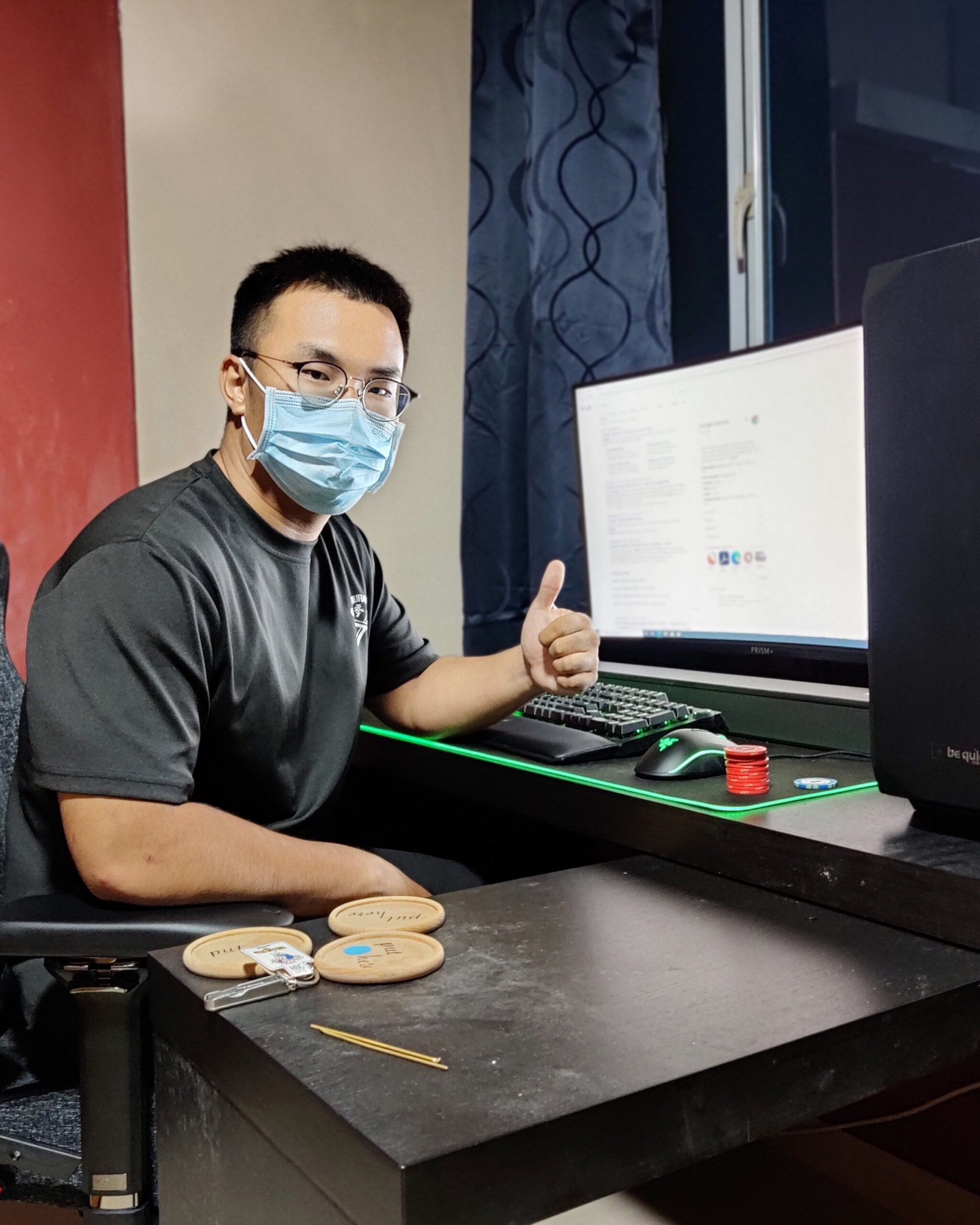 Satisfied PCExperts101 customer wearing a face mask and glasses, giving a thumbs up at a brightly lit computer desk, indicating successful PC service or repair.